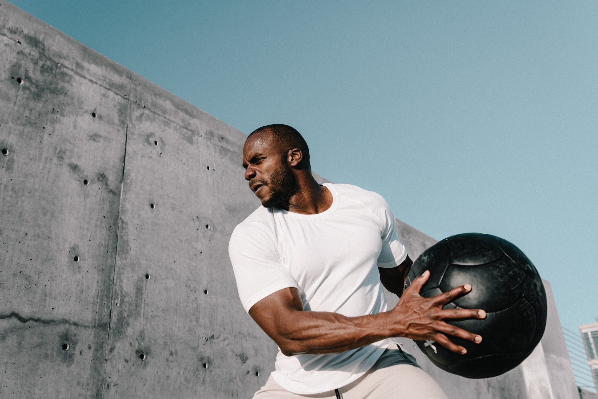 man working out with a large medicine ball outdoors, against a large concrete wall. 